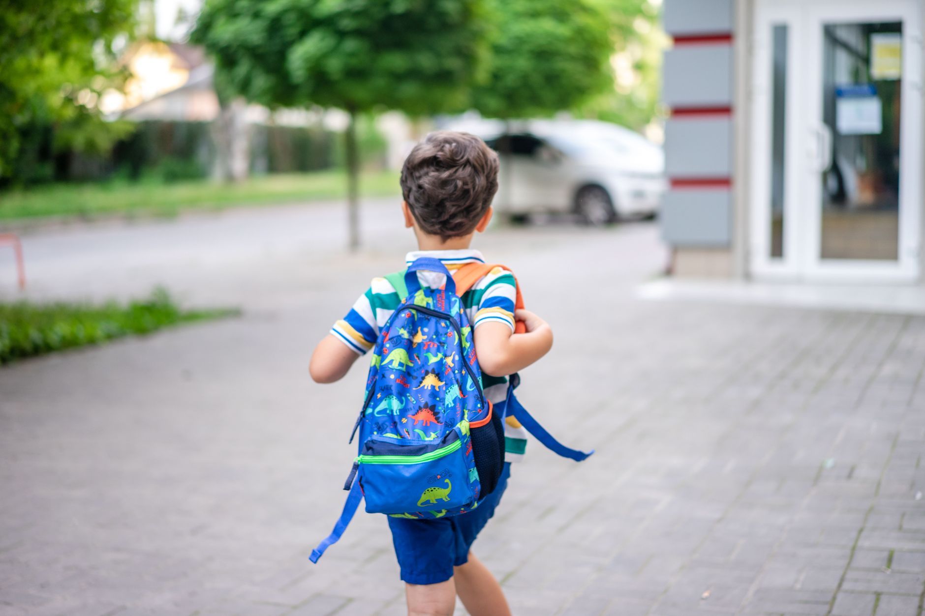 A young child with a backpack walking toward a school entrance on a summer morning