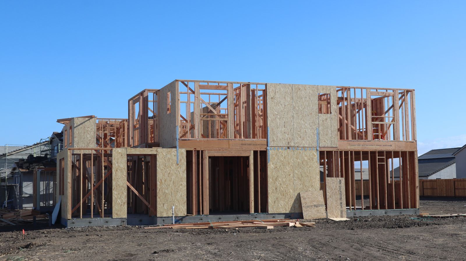 A two-storey wood-framed house under construction, partly sheathed, on a bare dirt lot beside finished homes under a clear blue sky