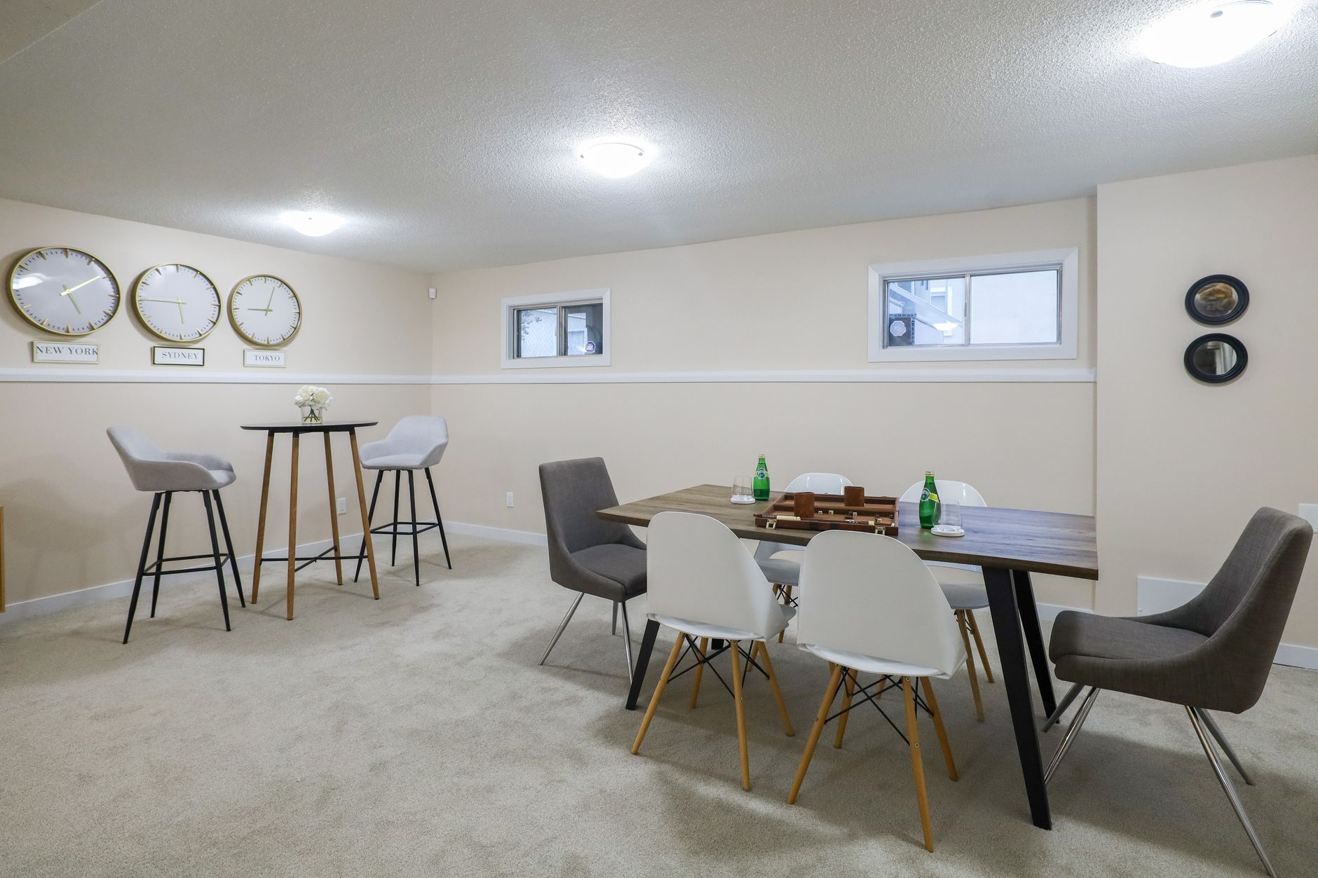A finished basement living area with pale carpet, a dining table and small windows set high in the wall