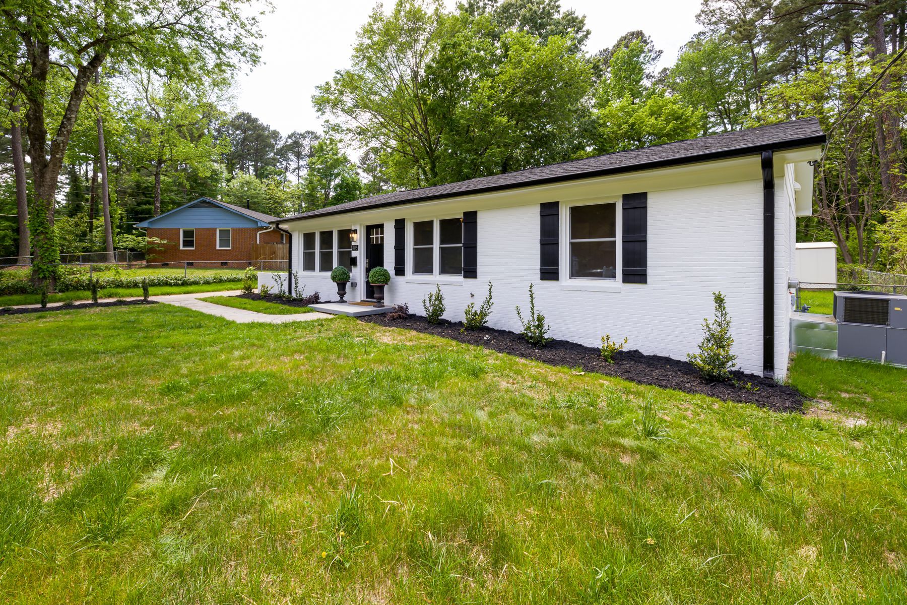 A single-storey white brick bungalow with black shutters set back behind a deep front lawn and mature trees