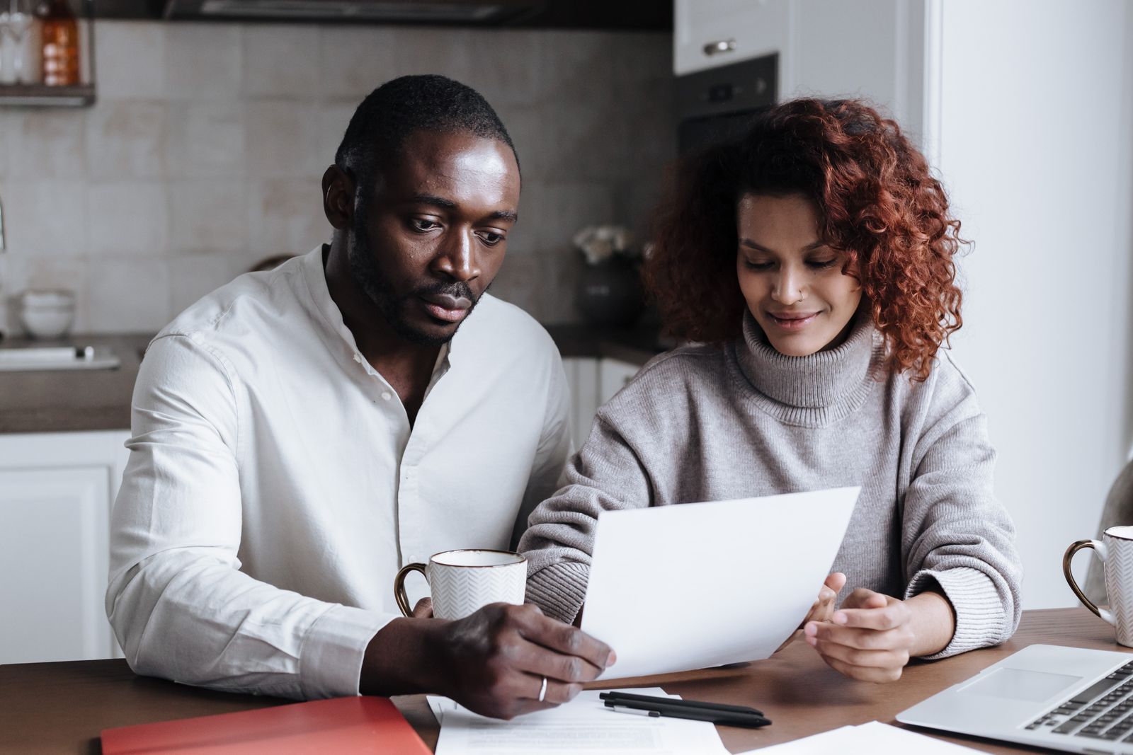 A couple at a kitchen table reading a printed document together, coffee mugs and an open laptop beside them