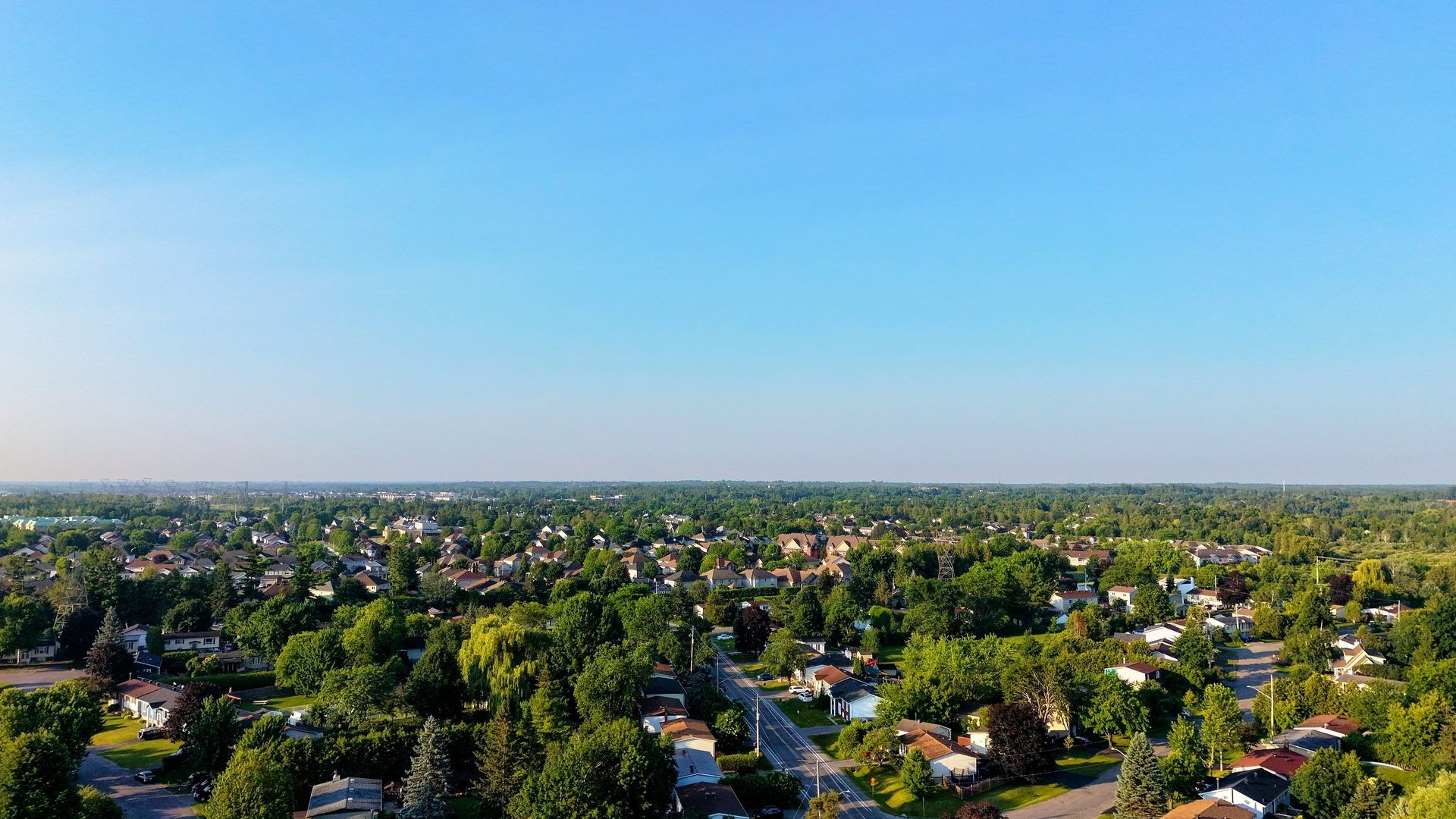 An aerial view across a residential neighbourhood of detached houses and mature trees under a clear blue sky