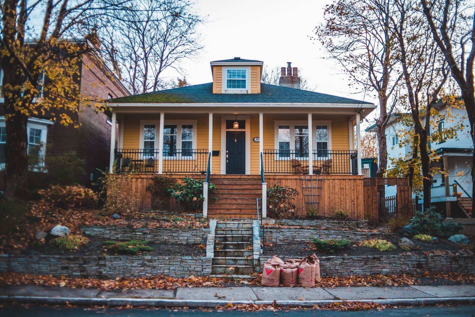A yellow bungalow with a covered front porch on a tree-lined street in late fall, bare branches overhead and paper leaf bags waiting at the curb