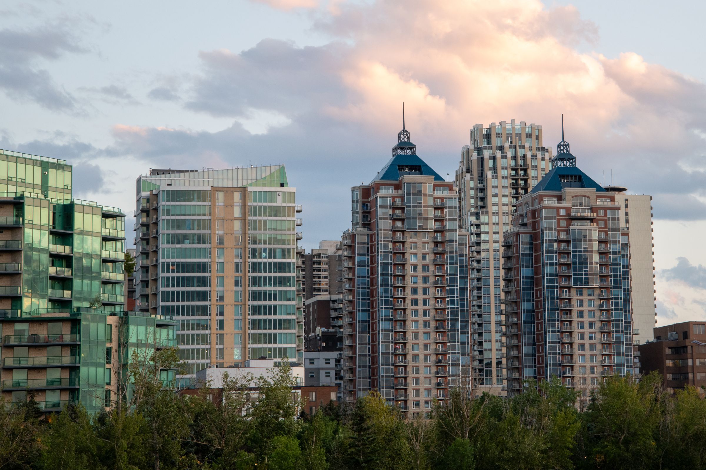 A cluster of Calgary residential condo towers from different eras, a green-glass modern building beside a beige mid-rise and brick-and-glass towers with blue peaked roofs, under a soft cloudy sky